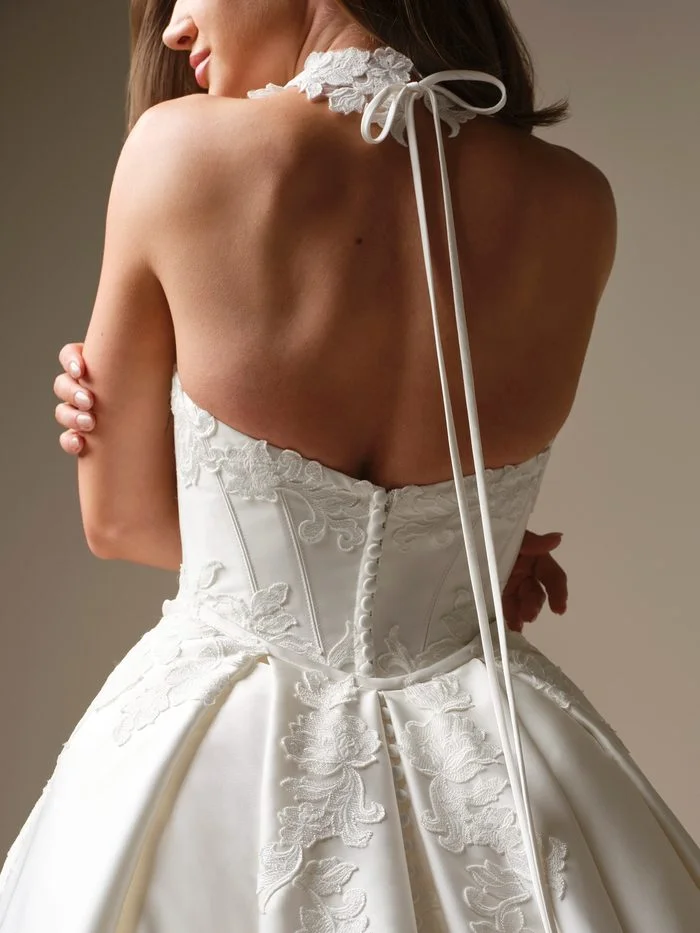 Back view of the Serafina Lane choker by Maggie Sottero, a band of embroidered floral lace tied at the nape with a thin satin ribbon whose long ends fall down the bride’s back to a covered-button ballgown with lace appliqués.