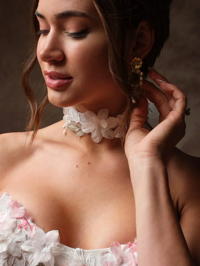Close-up of a bride touching her gold statement earring while wearing the Benton choker by Maggie Sottero, a band of soft white 3D petal flowers and silver embroidered leaves on sheer tulle. It complements the pink and white floral appliqués of the Benton ballgown.