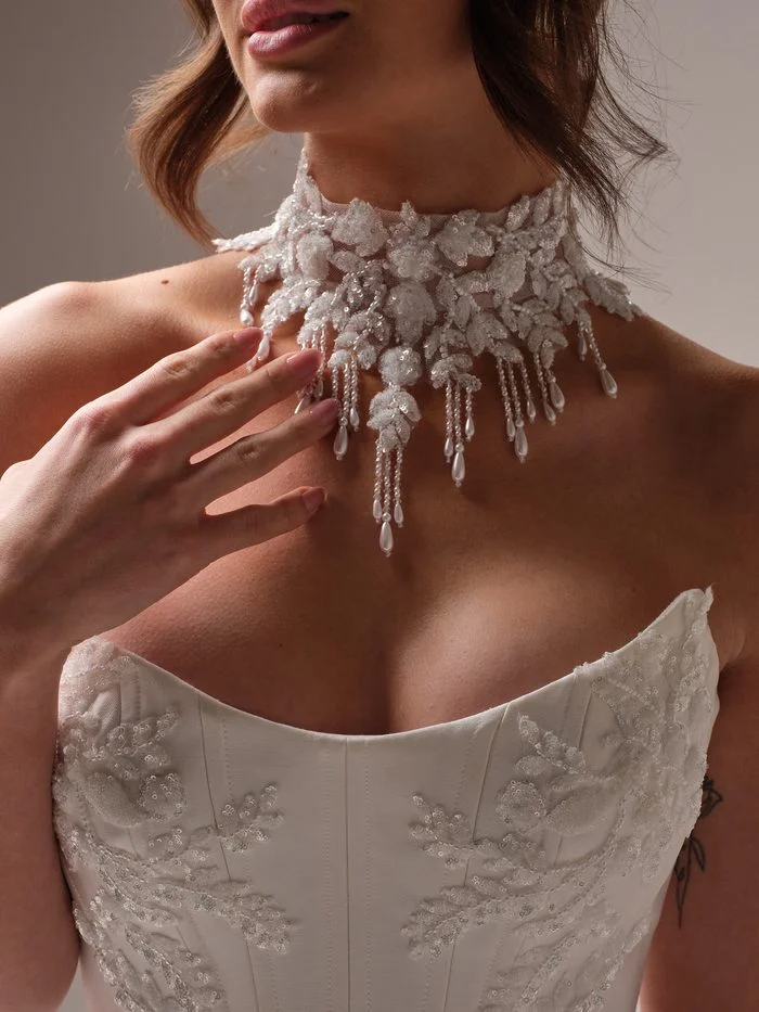 Close-up of the Amadora choker by Sottero and Midgley, with the bride’s hand resting below it. The high collar is covered in sequined and beaded floral leaves, with dangling strands of tiny beads ending in pearl teardrops above a beaded strapless corset.