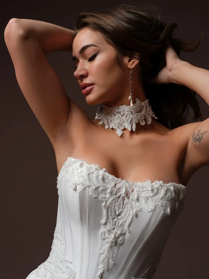 Bride with arms raised and hair swept back, wearing the Sandrine bridal choker by Sottero and Midgley, a wide collar of beaded floral lace with pearl and crystal accents. Long pearl drop earrings and the pearl-drop lace trim on the Sandrine ballgown’s structured corset complete the look.