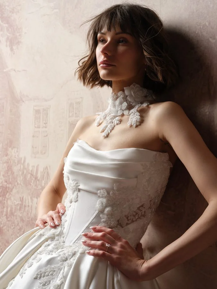 Bride with a tousled short bob and bangs leaning against a softly painted pink wall, wearing the Burke bridal choker by Sottero and Midgley. The high-neck choker is made of 3D lace flowers and sequined leaf sprays that extend down onto the collarbone, styled with the strapless draped satin Burke Lane ballgown.