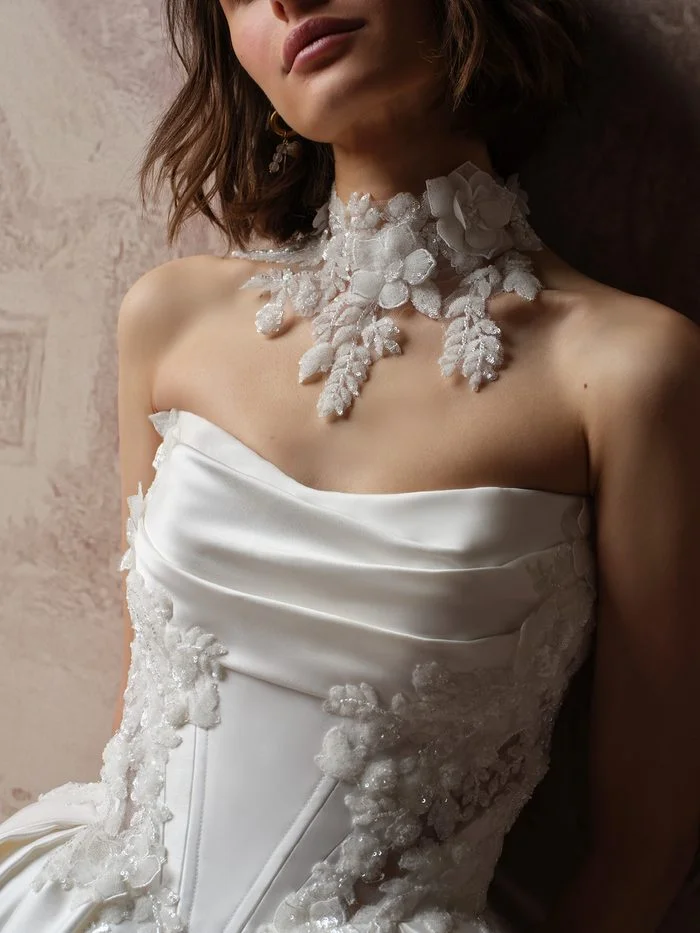 Close-up of the Burke bridal choker by Sottero and Midgley, showing its sheer high-neck base layered with dimensional white organza flowers and beaded, sequined leaves that fall in delicate sprays toward the neckline of a draped satin bodice.