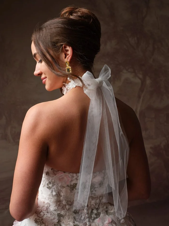 Back view of a bride with a sleek updo, showing the Benton wedding dress choker by Maggie Sottero fastened with an oversized sheer tulle bow. Long tulle streamers trail down her bare back toward the pink and sage floral appliqués of the gown.