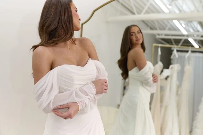 Bride tries on a chiffon wedding dress with a draped strapless neckline and sheer off-the-shoulder long sleeves, admiring the gown in a bridal boutique mirror.