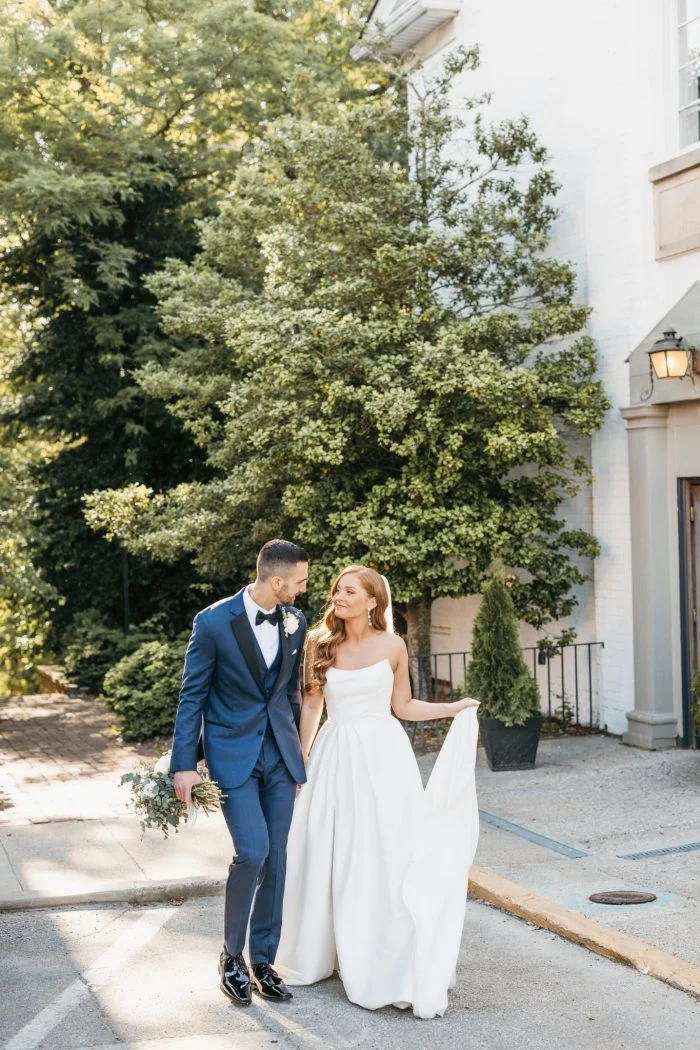 Bride and groom walk together outdoors, with the bride wearing a strapless Mikado wedding dress featuring a structured bodice and full A-line skirt.