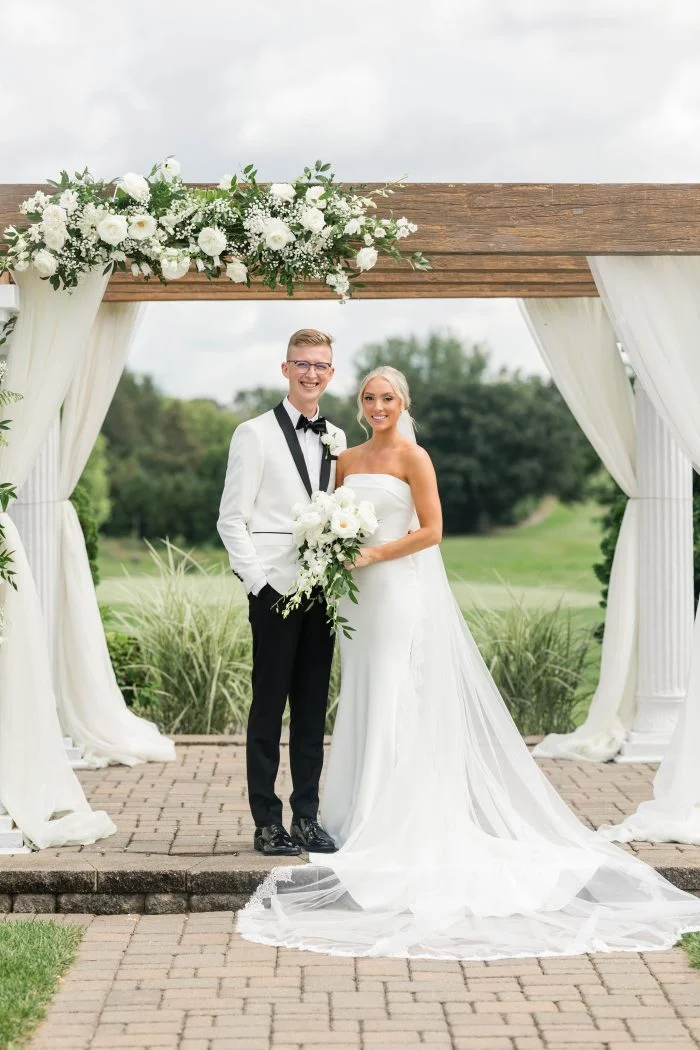 Bride and groom pose beneath a floral wedding arch, with the bride wearing a strapless crepe wedding dress with a flowing train.