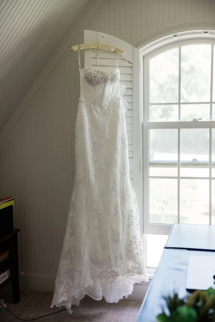 White floral lace wedding dress, Devon, hangs beside a sunlit window, featuring a sheer corset bodice, delicate spaghetti straps, draped pearl detailing, and a fitted silhouette with a flowing train.