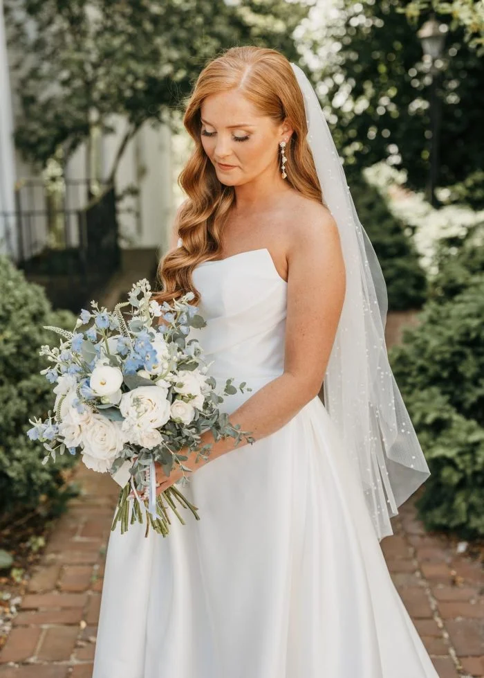 Bride in a strapless Mikado wedding dress with a draped neckline and flowing A-line skirt holds a bouquet of white and blue flowers, wearing a delicate pearl-dotted veil.