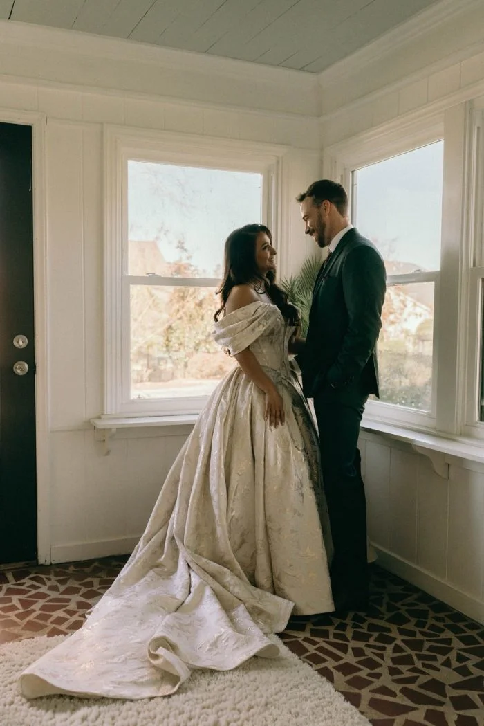 Bride and groom pose together by a window, with the bride wearing an off-the-shoulder floral jacquard ball gown wedding dress featuring a structured bodice, full skirt, and long train.