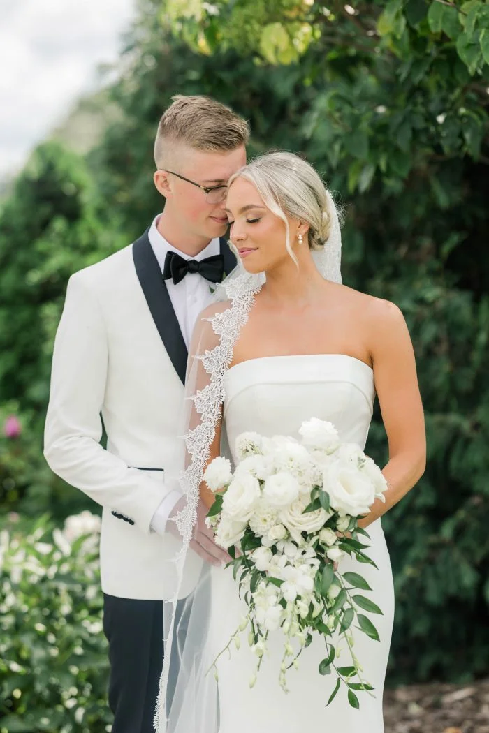 Close-up of a bride in a strapless crepe wedding dress with a lace-trimmed veil, holding a white floral bouquet beside the groom.