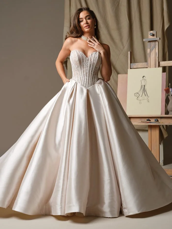 Ashland by Maggie Sottero in Blush, a ballgown wedding dress featuring a strapless sweetheart neckline, a corset-inspired lace bodice with visible boning, and a voluminous satin skirt with dramatic pleating. The gown is photographed in a fashion studio setting beside an artist’s easel and sketch canvas, highlighting the soft blush hue, structured silhouette, and elegant contrast between the intricate bodice and full skirt.