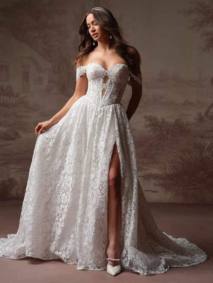 Narelle by Rebecca Ingram in Ivory over Misty Mauve, an A-line wedding dress featuring a sweetheart neckline, off-the-shoulder lace sleeves, and allover floral lace. The bodice showcases illusion cutout details at the center front, while the flowing A-line skirt features a thigh-high slit and sweeping train. Photographed in a studio setting, the gown highlights romantic floral texture and soft blush undertones beneath the ivory lace overlay.