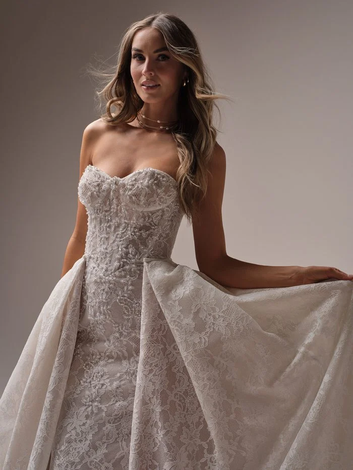 Jovienne by Sottero and Midgley in Ivory over Soft Blush, a fitted sheath wedding dress featuring a strapless sweetheart neckline, intricate lace appliqués, and pearl-accented embellishments throughout the bodice. A dramatic detachable lace overskirt adds volume and dimension, creating the look of a ballgown while showcasing the fitted silhouette beneath. The gown is photographed in a studio setting, highlighting the romantic lace texture and soft blush undertones.