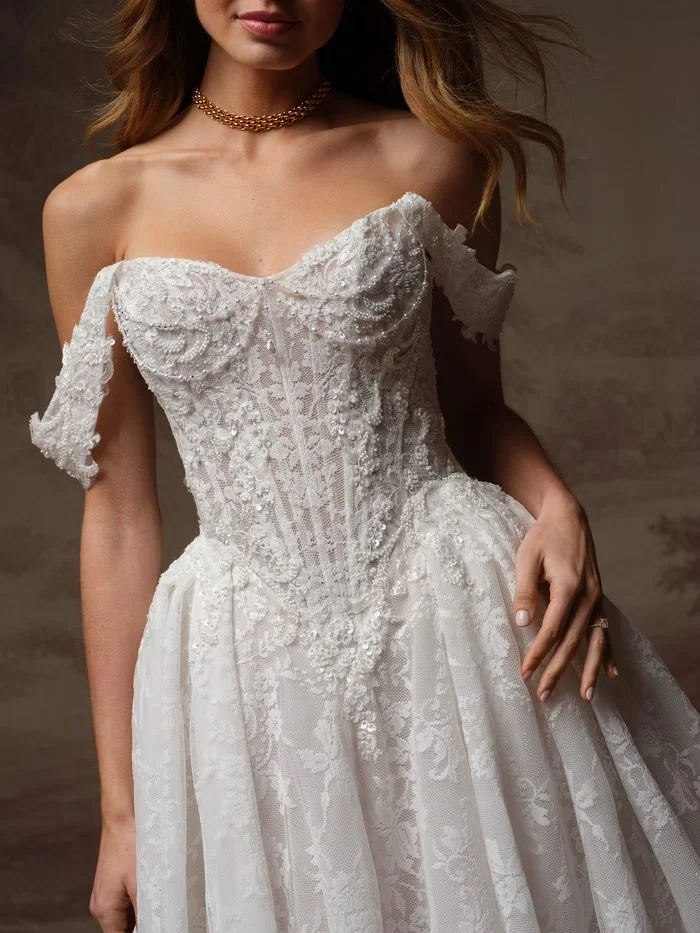 Close-up detail of Ryland by Maggie Sottero in Ivory over Blush, showcasing the sheer corset bodice with exposed boning, pearl-accented lace appliqués, and detachable off-the-shoulder lace sleeves. Intricate beading and dimensional floral embroidery add texture and depth, while the blush underlay creates soft contrast beneath the ivory lace overlay.