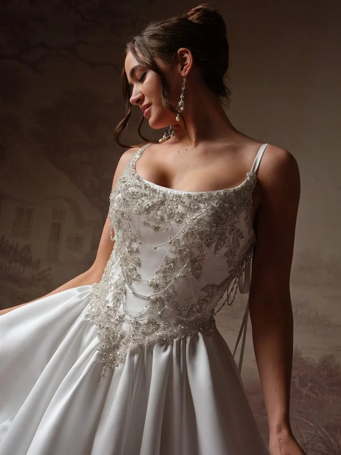 Aelarion by Sottero and Midgley, an A-line wedding dress featuring a scoop neckline, delicate shoulder straps with ribbon-tie accents, and intricate silver-beaded embellishments cascading across the bodice. The gown’s full satin skirt creates dramatic volume, while the detailed beading adds texture and shimmer. Photographed in a softly lit studio setting with a vintage-inspired backdrop.
