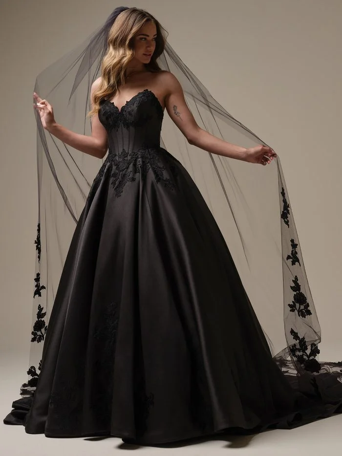 Serafina by Maggie Sottero in Black, a dramatic ballgown wedding dress featuring a strapless sweetheart neckline, a corset-inspired bodice with exposed boning, and matte black floral lace appliqués. The voluminous satin skirt creates a regal silhouette, while a coordinating black veil with embroidered floral accents frames the gown. Photographed in a studio setting, the design blends romantic detailing with gothic-inspired elegance.