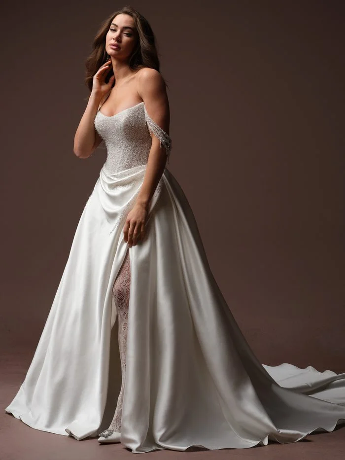 Rumi by Sottero and Midgley in Ivory, a ballgown wedding dress featuring a fully beaded bodice with shimmering silver crystal embellishments, a soft scoop neckline, and delicate off-the-shoulder beaded strands draping from the sleeves. A dramatic satin overskirt opens at the front to reveal a fitted lace silhouette beneath, creating a striking layered effect. The gown is photographed in a studio setting with a flowing chapel-length train that highlights its glamorous, couture-inspired design.