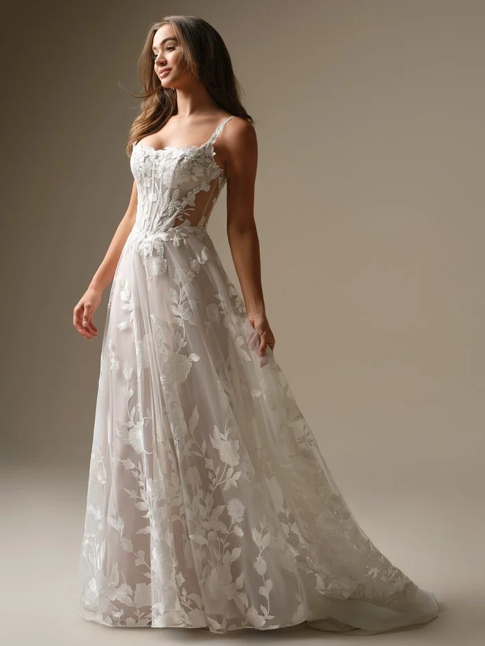 Natalie Marie by Maggie Sottero in Ivory over Blush, an A-line wedding dress featuring delicate spaghetti straps, a sheer corset-inspired bodice with exposed boning, and oversized floral lace appliqués cascading throughout the skirt. The full-length gown is shown in a softly lit studio setting, highlighting the romantic blush underlay, airy tulle layers, and sweeping train.