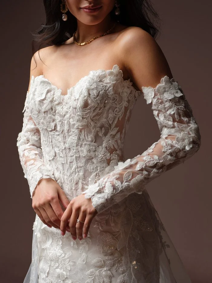Detachable Estelle wedding dress sleeves by Maggie Sottero in Ivory, featuring sheer illusion long sleeves adorned with dimensional floral lace appliqués and delicate beaded accents. The close-up image highlights the off-the-shoulder design, intricate floral textures, and coordinated embellishments that complement the Estelle fit-and-flare wedding gown.