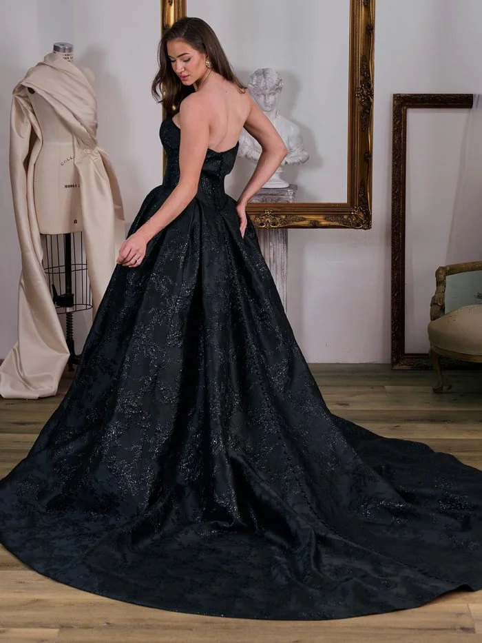 Aberdeen by Sottero and Midgley in Black, a dramatic ballgown wedding dress featuring a strapless sweetheart neckline, a structured corset bodice, and a full skirt crafted from black floral jacquard fabric with a subtle tonal shimmer. The voluminous silhouette extends into a sweeping train, creating a regal statement. Photographed in a bridal atelier setting with ornate gold frames and vintage furnishings, the gown showcases modern couture styling with gothic-inspired elegance.