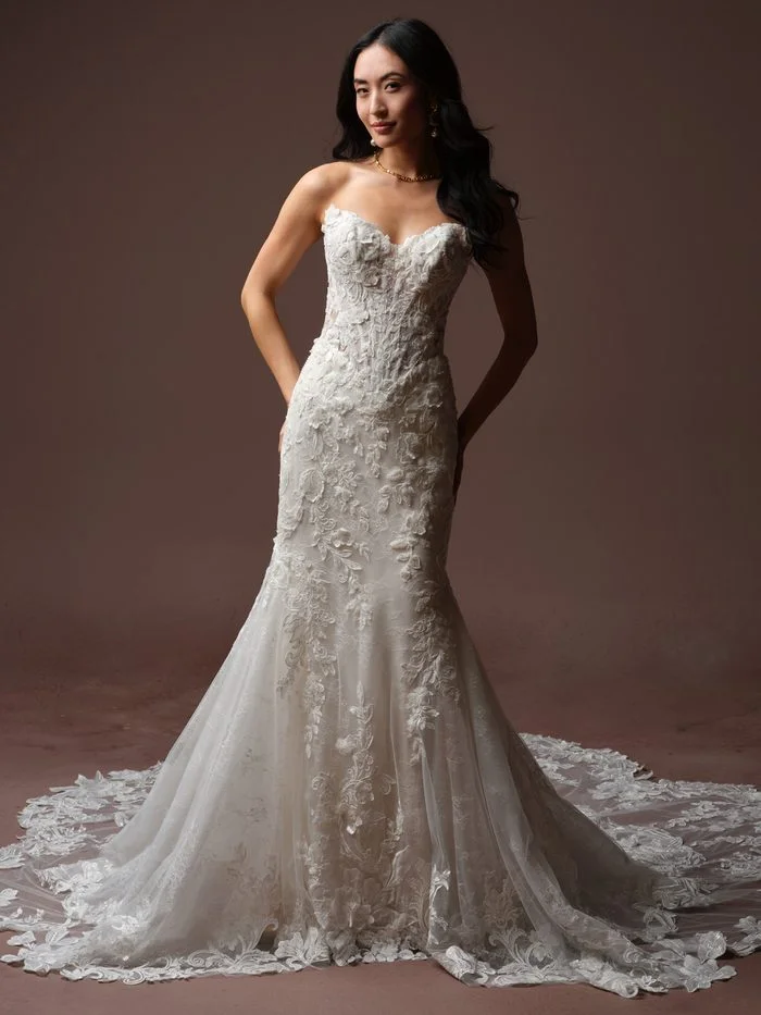 Estelle by Maggie Sottero in Ivory over Misty Mauve, a fit-and-flare wedding dress featuring a sweetheart neckline, sheer illusion side panels, and dimensional floral lace appliqués accented with subtle beading. The fitted silhouette highlights intricate botanical embroidery that flows into a soft tulle skirt and scalloped lace train. Photographed in a studio setting, the gown showcases richly textured floral details and romantic couture-inspired craftsmanship.