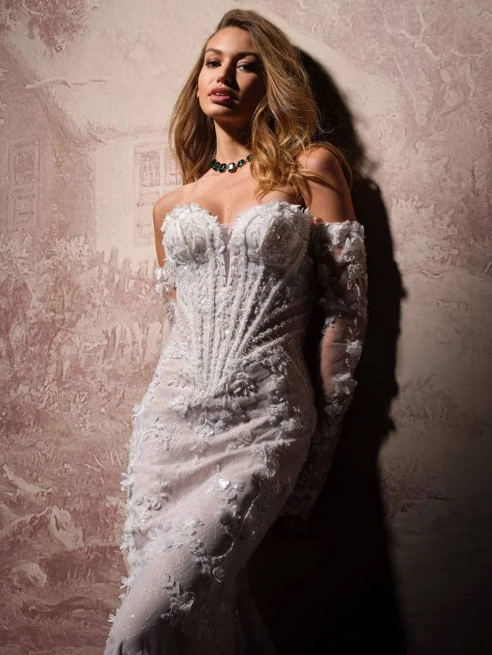 Stasia by Maggie Sottero in Ivory over Blush, a fitted wedding dress featuring a structured corset bodice with a plunging illusion neckline, dimensional floral lace appliqués, and off-the-shoulder illusion sleeves. The gown is shown in a close-up portrait highlighting the intricate beading, textured lace details, and figure-hugging silhouette against a softly lit vintage-inspired backdrop.