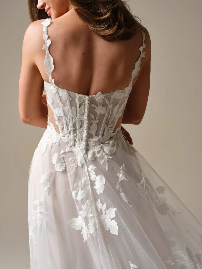 Back view of Natalie Marie by Maggie Sottero in Ivory over Blush, an A-line wedding dress featuring delicate floral lace straps, a sheer illusion bodice with exposed corset boning, and a low scoop back accented with covered buttons. Oversized floral lace appliqués frame the waist and cascade onto the flowing tulle skirt, while the blush underlay adds soft dimension beneath the ivory lace overlay. The gown is photographed in a clean studio setting, highlighting the intricate back detailing and airy silhouette.