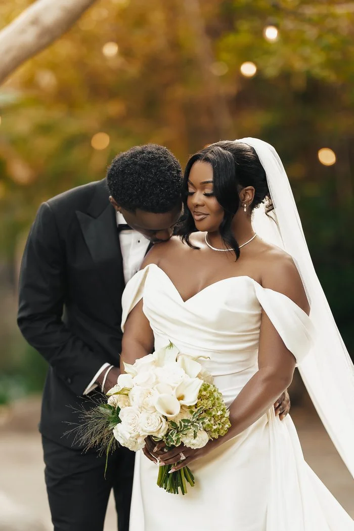 Bride wearing the Sottero and Midgley Giselle wedding dress, an ivory fitted gown with an off-the-shoulder draped neckline and softly pleated bodice. The bride holds a bouquet of white flowers while standing with a partner in a black tuxedo beneath softly glowing lights. Photo by JAO Photography