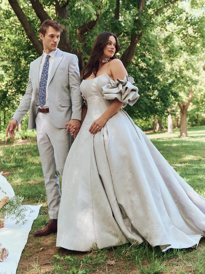 Outdoor lifestyle image of a model wearing the Maggie Sottero Fortune Marie ballgown wedding dress. The gown features a straight strapless neckline, a structured bodice, and a full jacquard ballgown skirt. Voluminous detachable puff sleeves add a dramatic statement as the model walks through a park setting beside a partner in a light-colored suit.