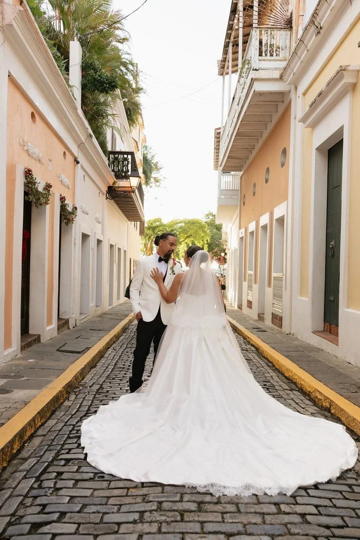 Back view of a bride wearing the Maggie Sottero Serafina wedding dress standing with a partner on a cobblestone street lined with colorful historic buildings. The ivory ballgown features a full skirt, chapel-length train, covered buttons down the back, and a lace-trimmed veil cascading over the train. Photo by Hector Javier.