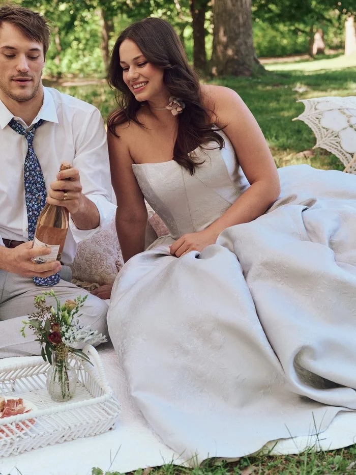Outdoor lifestyle image of a model wearing the Maggie Sottero Fortune Marie ballgown wedding dress while seated on a picnic blanket. The strapless jacquard ballgown features a structured bodice and full skirt with subtle floral texture. A floral choker accessory completes the romantic garden-inspired look.