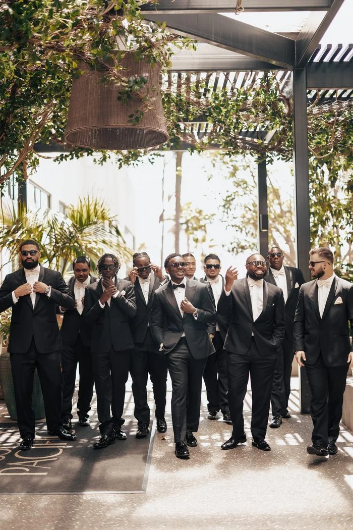 Group portrait of a wedding party beneath a vine-covered pergola. The groom and groomsmen wear classic black tuxedos with bow ties and sunglasses, walking toward the camera in an outdoor venue. Photo by JAO Photography.
