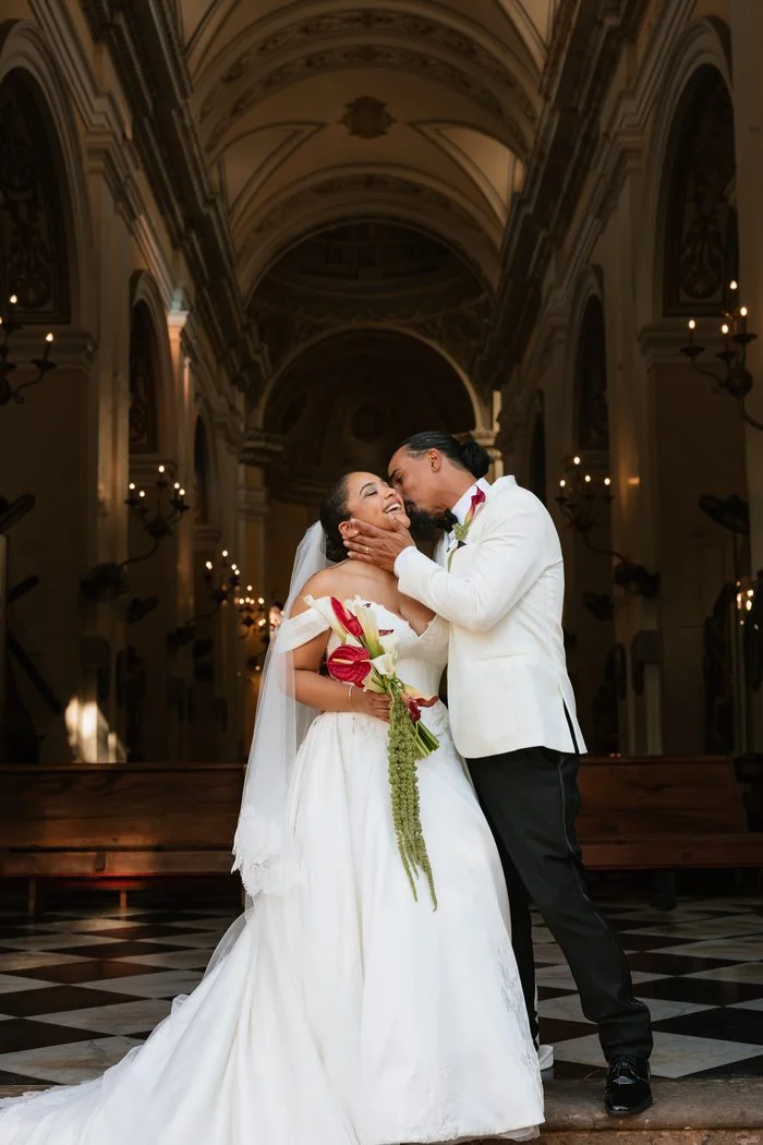 Bride wearing the Maggie Sottero Serafina wedding dress posing with a partner inside a cathedral. The ivory ballgown features an off-the-shoulder neckline, lace-accented bodice, and full skirt with a sweeping train. The bride carries a bouquet of white calla lilies, red anthuriums, and cascading greenery. Photo by Hector Javier.