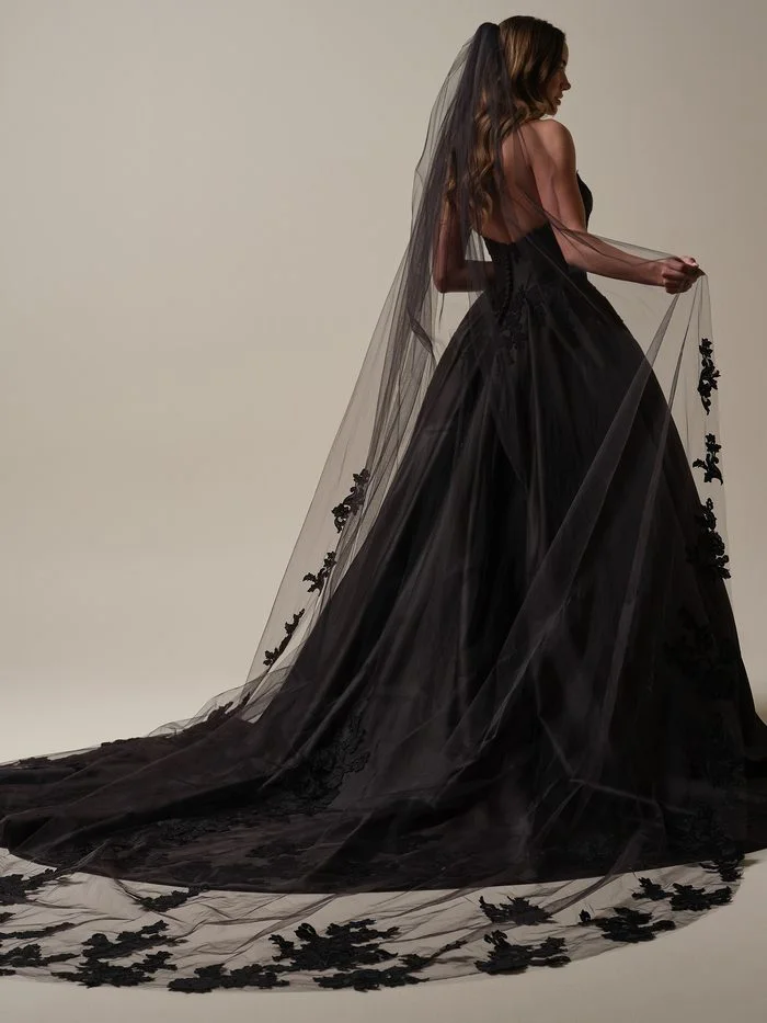 Back view of a model wearing the Maggie Sottero Serafina ballgown wedding dress in black. The strapless corset bodice reveals an open back, while the voluminous satin ballgown skirt extends into a dramatic train. A black cathedral-length veil with floral lace appliqués cascades behind the gown and pools along the floor.