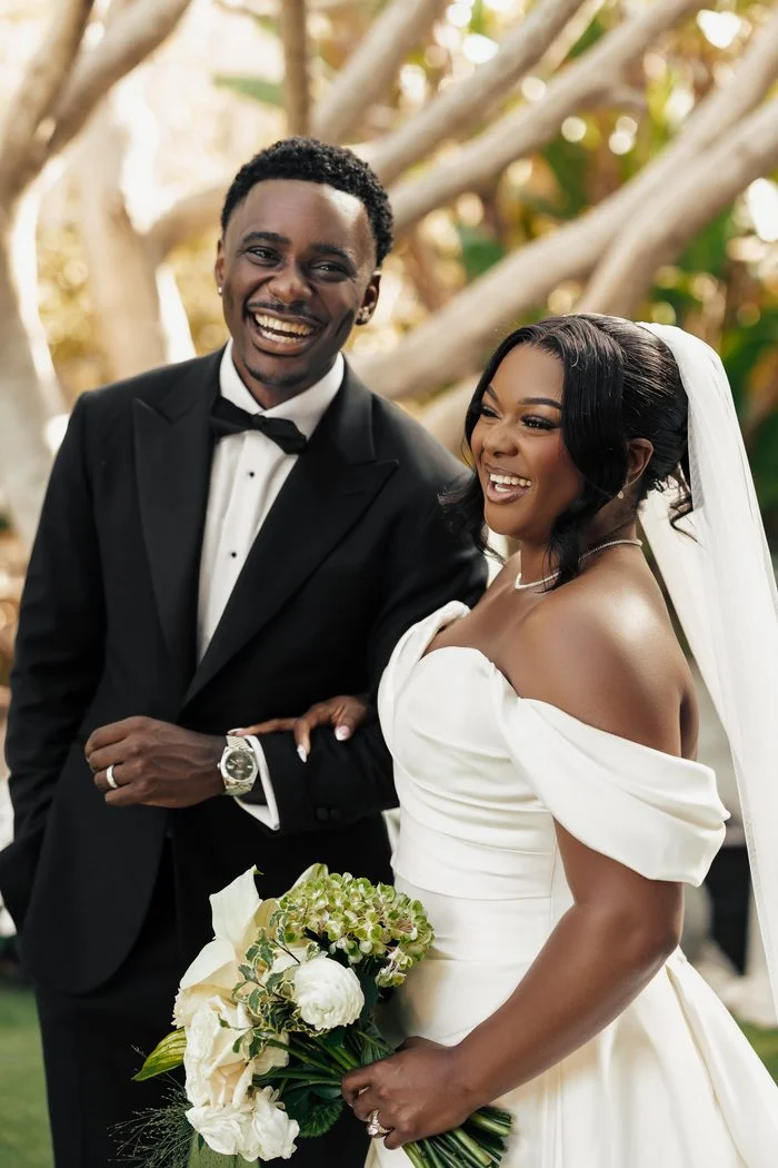 Bride wearing the Sottero and Midgley Giselle wedding dress standing arm-in-arm with a partner in a black tuxedo. The ivory gown features an off-the-shoulder draped neckline, softly pleated bodice, and fitted silhouette. The bride carries a bouquet of white roses, calla lilies, and green hydrangeas, with a chapel-length veil completing the look. Photo by JAO Photography.