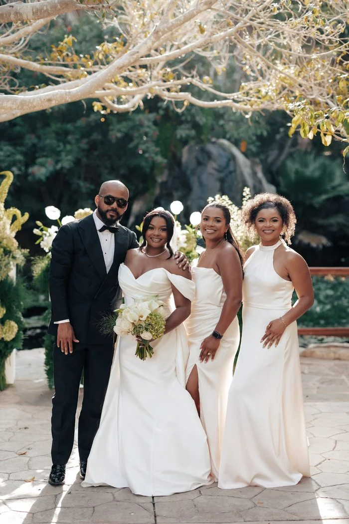 Bride wearing the Sottero and Midgley Giselle wedding dress posing with a man in a tux and bridal wedding party in a garden setting. The ivory gown features an off-the-shoulder neckline, draped bodice, and fitted silhouette. The group stands beneath sunlit tree branches surrounded by lush greenery. Photo by JAO Photography.