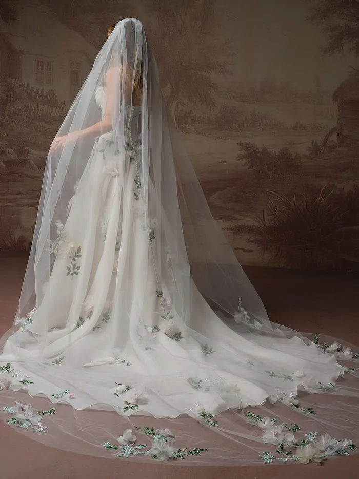 Back view of a model wearing the Sottero and Midgley Amaryllis wedding dress with the Amaryllis veil. The ivory A-line gown features three-dimensional floral appliqués with subtle pastel accents, while the cathedral-length tulle veil cascades over the gown and train, finished with scattered floral embellishments along the edge.