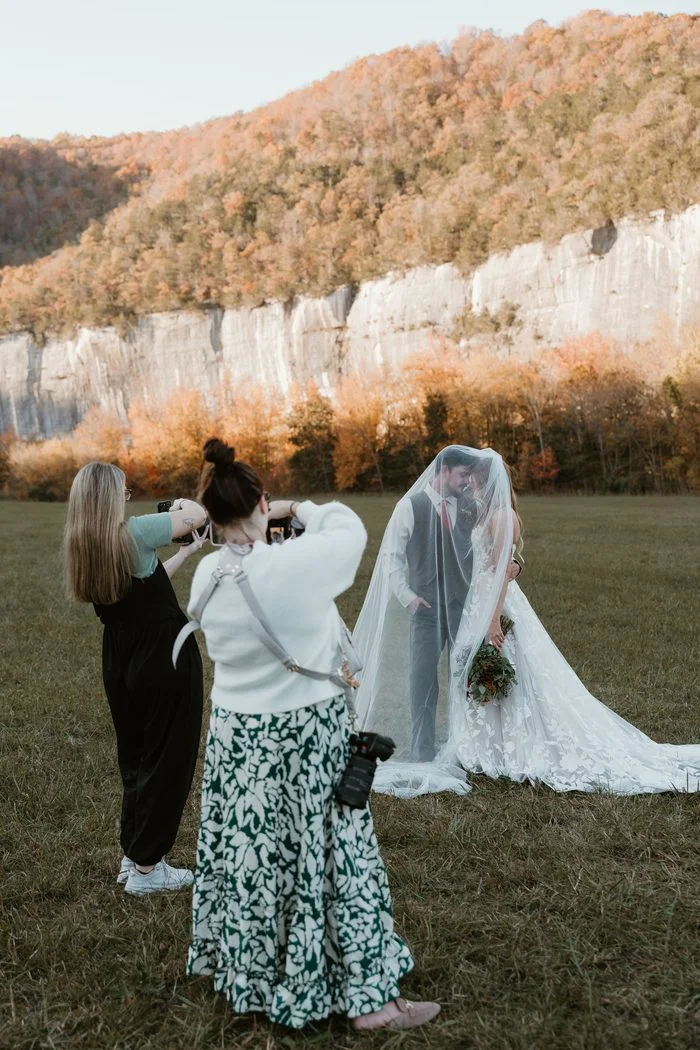 Behind-the-scenes photo of two photographers capturing a couple standing beneath a cathedral-length veil in an open field. The bride wears Natalie by Maggie Sottero, an ivory A-line wedding dress featuring a strapless floral lace bodice, sheer corset detailing, and oversized botanical lace motifs cascading throughout the skirt and train. Limestone cliffs and autumn foliage create a dramatic natural backdrop.