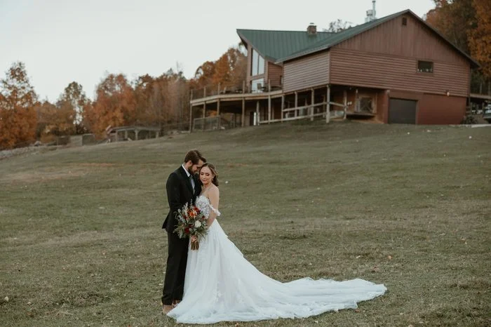 Couple posing on a grassy hillside with a rustic lodge and autumn trees in the background. The bride wears Wicklow by Sottero and Midgley, an ivory A-line wedding dress featuring an off-the-shoulder floral lace bodice, dimensional lace appliqués, and a flowing tulle skirt with a sweeping train. The bride holds a bouquet of rust, ivory, and greenery florals while standing beside a suited partner.