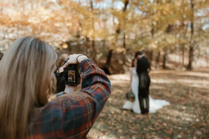 Behind-the-scenes photo of a photographer capturing a couple in an autumn woodland setting. The bride wears Maggie Sottero, an ivory A-line wedding dress featuring a strapless floral lace bodice, sheer corset detailing, and oversized botanical lace motifs throughout the skirt and train. Fallen leaves blanket the forest floor while the couple poses in the distance among sunlit trees.