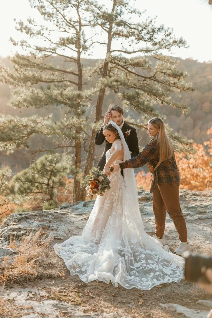 Behind-the-scenes photo of a stylist adjusting a bride’s bouquet and gown on a rocky overlook. The bride wears Natalie by Maggie Sottero, an ivory A-line wedding dress with a strapless floral lace bodice, sheer corset construction, and oversized botanical lace appliqués cascading across the skirt and sweeping train. Pine trees, golden autumn foliage, and warm sunlight frame the outdoor portrait setting.