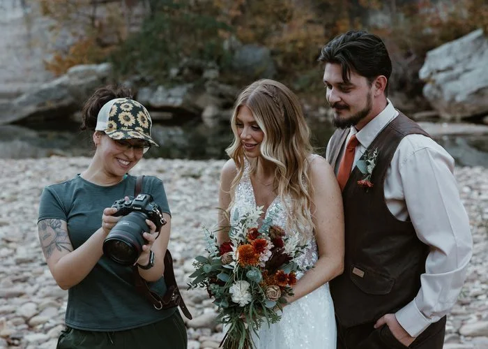 Behind-the-scenes photo of a photographer reviewing images on a camera with a couple standing beside a rocky riverside. The bride wears an ivory Maggie Sottero wedding dress featuring a fitted embellished bodice, shimmering glitter tulle skirt, and delicate floral details. The bride holds a bouquet of rust, burgundy, ivory, and greenery florals while the couple looks at the camera display.