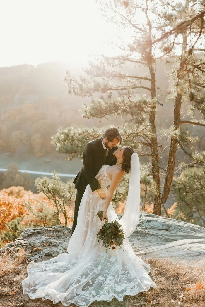 Couple posing on a rocky overlook surrounded by pine trees and autumn foliage. The bride wears Natalie by Maggie Sottero, an ivory A-line wedding dress featuring a strapless floral lace bodice, sheer corset detailing, and oversized botanical lace motifs throughout the skirt and train. A fingertip veil and a bouquet of rust, ivory, and greenery florals complete the look as the couple stands against a sunlit landscape backdrop.