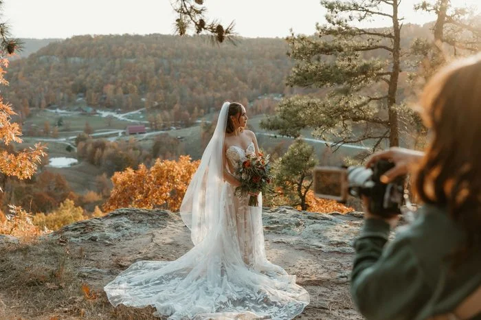 Behind-the-scenes photo of a photographer capturing a bridal portrait on a scenic overlook above a valley of autumn trees. The bride wears Natalie by Maggie Sottero, an ivory A-line wedding dress with a strapless floral lace bodice, sheer corset construction, and oversized botanical lace appliqués cascading across the skirt and sweeping train. A pearl-detailed veil trails behind the bride as warm golden light illuminates the landscape.