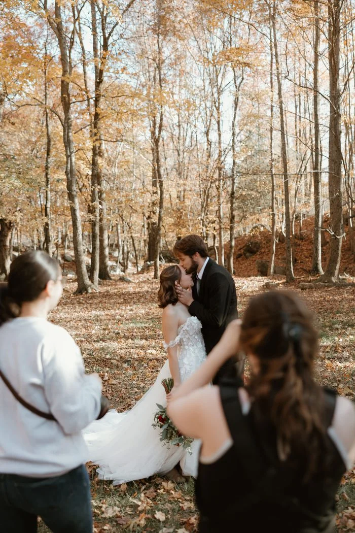Behind-the-scenes photo of photographers capturing a couple kissing in an autumn woodland clearing. The bride wears Wicklow by Sottero and Midgley, an ivory A-line wedding dress featuring an off-the-shoulder floral lace bodice, dimensional lace appliqués, and a voluminous tulle skirt that extends into a flowing train. Golden foliage and fallen leaves create a warm seasonal backdrop.