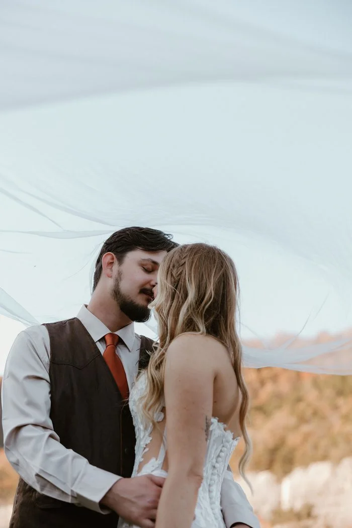 Close-up of a couple standing beneath a billowing cathedral-length veil outdoors. The bride wears Natalie by Maggie Sottero, an ivory A-line wedding dress with a strapless floral lace bodice, sheer side panels, and a structured corset back adorned with oversized botanical lace appliqués. Warm autumn colors and towering limestone cliffs frame the romantic portrait.