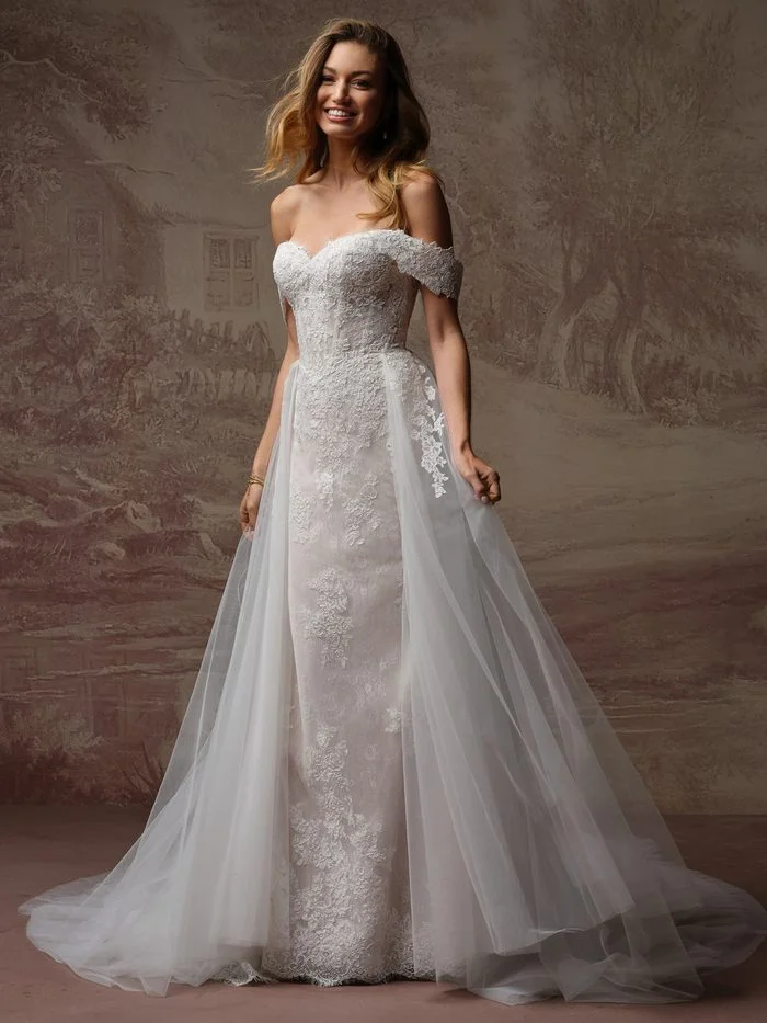 Rebecca Ingram Esmeralda Lane fit-and-flare wedding gown featuring floral lace, an off-the-shoulder neckline, and a detachable tulle overskirt for petite brides.