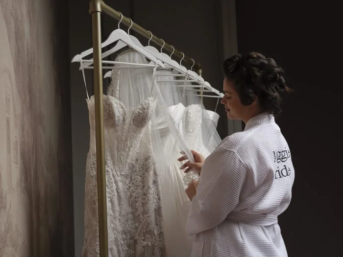 Bride getting ready in a robe while looking through a rack of lace wedding dresses and bridal veils.