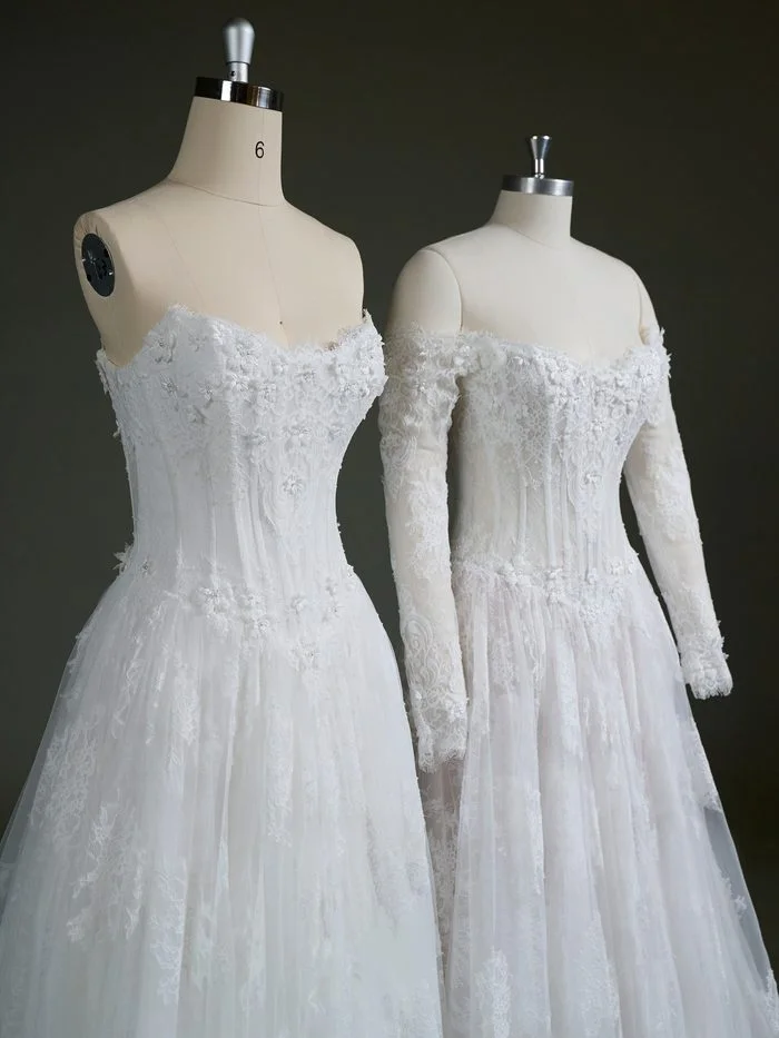 Close-up view of two Kitaro by Maggie Sottero wedding gowns displayed on dress forms. Both feature a lace-covered corset bodice with floral appliqués, delicate beadwork, and a sweetheart neckline. The left gown showcases the strapless design, while the right gown is styled with detachable off-the-shoulder long sleeves made from matching lace, highlighting the gown’s customizable sleeve option.