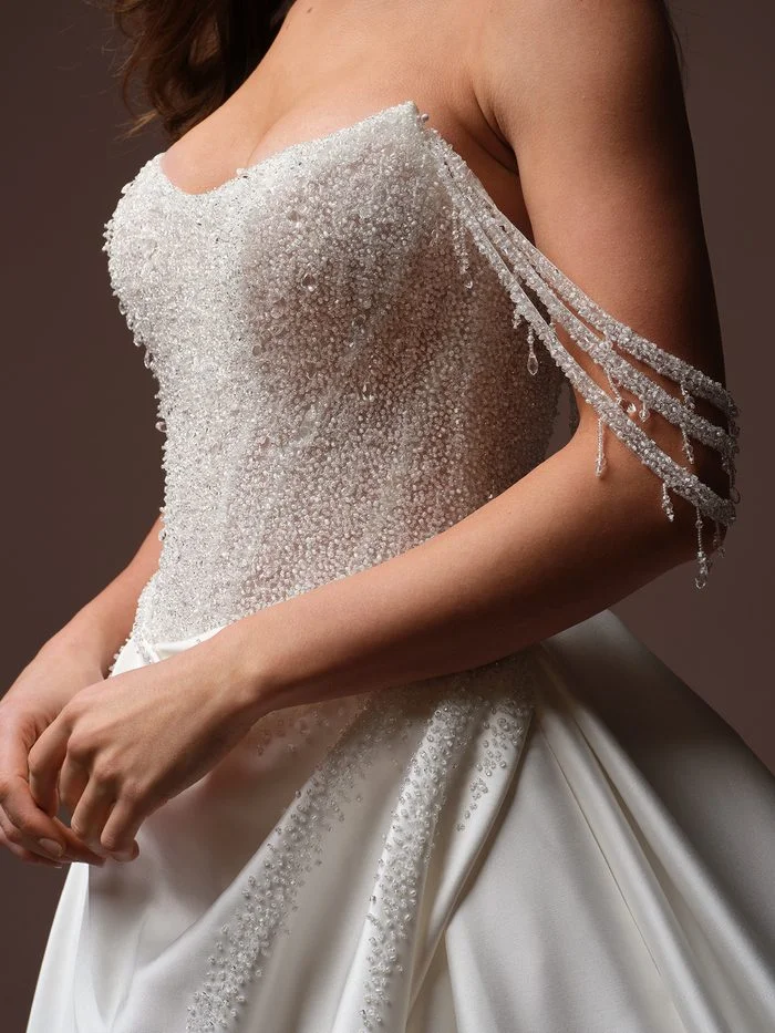 Close-up of the Rumi by Sottero and Midgley wedding gown featuring a strapless sweetheart neckline and a couture-inspired ballgown silhouette. The bodice is densely embellished with shimmering beadwork, crystals, and sequins, while detachable off-the-shoulder beaded sleeves drape gracefully along the upper arms with crystal drop accents. The satin skirt is finished with scattered beaded detailing that echoes the sparkle of the bodice and sleeves.